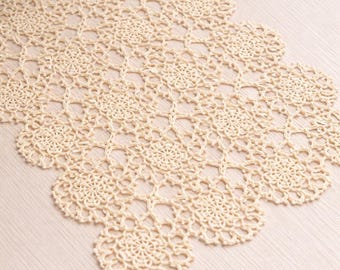 May include: A cream-colored, crocheted table runner with a floral pattern. The runner is made of a delicate, open-weave material and is placed on a light-colored wooden surface. The runner's edges have a scalloped design.