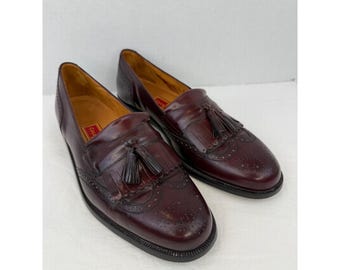 May include: A pair of burgundy leather loafers with decorative perforations and tassel accents. The shoes have a classic design with a rounded toe and a black sole. The loafers are displayed on a white surface.