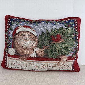 May include: Rectangular tapestry pillow with a red border. The image features a fluffy cat wearing a Santa hat, holding a Christmas tree, and a red bird. The text "KITTY KLAUS" is at the bottom.