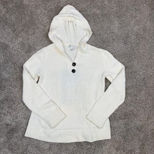 May include: A cream-colored knit hooded sweater featuring a two-button placket, long sleeves with ribbed cuffs, and a front kangaroo pocket. The garment is constructed with a vertical cable knit pattern and a solid ribbed hem at the bottom.