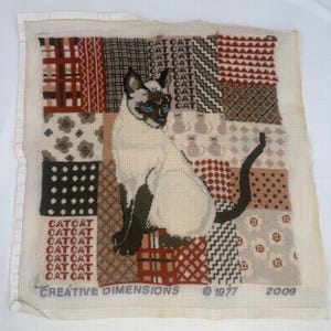 May include: A square needlepoint canvas featuring a central Siamese cat with blue eyes, surrounded by a patchwork of geometric, floral, and polka dot patterns. The text CAT repeats in columns, and the bottom reads CREATIVE DIMENSIONS 1977 2009.
