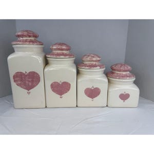 Może przedstawiać: Zestaw czterech ceramicznych pojemników o różnych rozmiarach. Każdy pojemnik jest w kolorze złamanej bieli z różowym wzorem serca i pasującą różową pokrywką. Pojemniki są ustawione w rzędzie, prezentując swoje różne wysokości i kształty.
