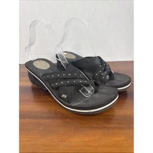 May include: A pair of black Skechers sandals with white accents. The sandals feature a thong-style design with adjustable straps and small white dot embellishments. The soles have a white stripe and a small wedge heel.