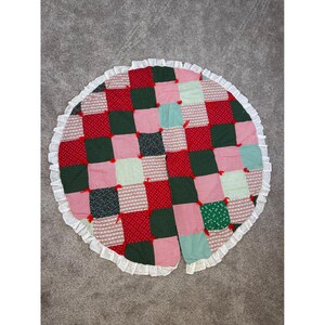 May include: A round, patchwork tree skirt with a white lace edge. The skirt features a checkerboard pattern of red, green, pink, and light blue squares. Some squares have a dotted or striped design. The skirt is designed to go around a Christmas tree.