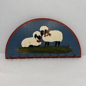 May include: A semi-circular wooden wall rack featuring a hand-painted scene of two white sheep on a blue background. The standing sheep wears a red polka-dot bow, while the sitting sheep has a red heart. Nine small wooden pegs line the bottom edge.
