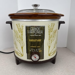 Może przedstawiać: Zabytkowy wolnowar Crock Watcher firmy Hamilton Beach. Urządzenie ma kremowy wygląd z brązowymi akcentami i szklaną pokrywą. Posiada wyjmowany pojemnik o pojemności 3,8 litra i funkcję automatycznej zmiany.