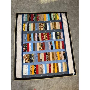 Op de afbeelding: Een rechthoekige patchworkquilt met een zwarte rand en een beige rand. De quilt heeft verticale stroken van verschillende stoffen met patronen in kleuren als rood, blauw, bruin en geel, gescheiden door lichtblauwe banden.