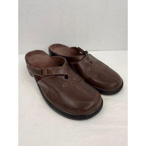 May include: A pair of brown leather clogs with adjustable straps. The clogs have a rounded toe, decorative stitching, and a black sole. The shoes appear to be unworn and are set against a white background.