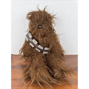Op de afbeelding: Een pluche Chewbacca-pop, met harige bruine vacht en een bandolier met zilveren en bruine details. De pop heeft een zwarte neus en zit op een houten oppervlak. Een Star Wars-verzamelobject.