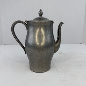 May include: A metallic gray pewter coffee pot with a curved handle, a long spout, and a removable lid featuring a small finial knob. The surface of the vessel has a matte, slightly weathered finish with visible signs of age and use throughout.