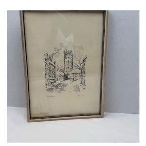 May include: A framed black and white ink sketch on off-white paper depicting a European town square with a central stone tower, surrounding buildings, and bare trees. The artwork is enclosed in a simple light-colored wooden frame with glass.