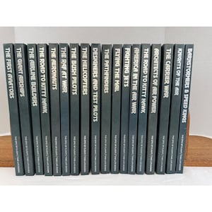 May include: A collection of vintage books with dark gray covers, featuring titles like "The First Aviators" and "Barnstormers & Speed Kings." Each book has white text and is part of "The Epic of Flight / Time-Life Books" series. The books are arranged on a wooden surface.