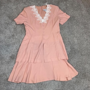 May include: A dusty rose dress featuring short sleeves, a V-neckline with white floral lace trim, and a front button closure. The garment has a layered skirt design with a split front panel and a flared hemline, providing a structured silhouette.