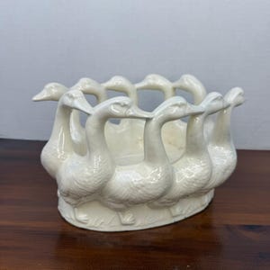 Może przedstawiać: Kremowa ceramiczna miska z ciągłym pierścieniem wyrzeźbionych gęsi tworzących zewnętrzną ściankę. Gęsi przedstawiono w pozycji stojącej z szyjami wygiętymi do wewnątrz, tworząc okrągłą konstrukcję o gładkim, błyszczącym wykończeniu powierzchni.