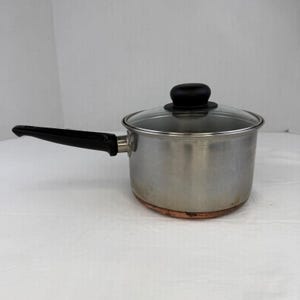 May include: A stainless steel saucepan with a copper-coloured base, a long black handle, and a clear glass lid featuring a black knob. The cooking pot has a cylindrical body and a polished metallic finish, designed for stovetop use in a kitchen.