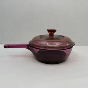 May include: A translucent purple glass saucepan with a matching lid. The pan features a long, flat handle extending from one side and a circular lid with a central knob. The glass has a deep violet hue and a smooth, polished finish throughout.