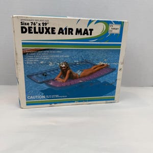 Puede incluir: Una caja de cartón rectangular para un colchón inflable Deluxe Air Mat con fondo blanco, detalles gráficos en azul y verde y texto en negro. La caja muestra el texto Size 76 inches by 29 inches y una imagen de un colchón inflable morado.
