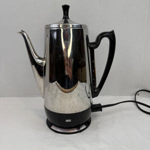 Könnte beinhalten: Eine elektrische Perkolator-Kaffeemaschine aus poliertem Edelstahl mit schwarzem Kunststoffgriff, Sockel und Deckelknauf. Der Griff verfügt über ein vertikales Sichtfenster mit Markierungen für 4, 6, 8 und 10 Tassen. Ein schwarzes Stromkabel.