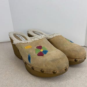 May include: A pair of tan suede clogs with a floral embroidery design in yellow, green, blue, and pink. The clogs have a wooden sole with brass studs and a faux fur lining.