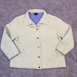 May include: A light yellow quilted jacket featuring a collared neckline, long sleeves, and a front button closure. The interior lining is a contrasting light purple color with a quilted texture. The jacket includes two front pockets.