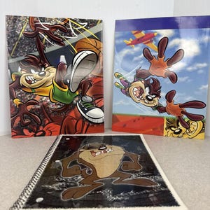 May include: Three notebooks featuring the Tasmanian Devil cartoon character. One notebook shows the character playing basketball, another shows him flying with a plane, and the third is a spiral-bound notebook with the character on the cover.