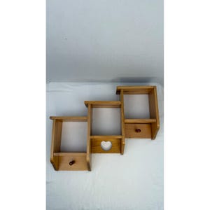 May include: A tiered wooden wall shelf unit featuring three connected rectangular compartments. The two lower shelves each include a small round wooden knob, and the middle shelf section displays a heart-shaped cutout in the lower panel.