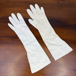 Op de afbeelding: Een paar lange, gebroken witte handschoenen gemaakt van een gladde, lichtgewicht stof. Deze operalange handschoenen reiken tot voorbij de pols en hebben een eenvoudig ontwerp met gestikte vingernaden en een rechte opening bij de onderarm.