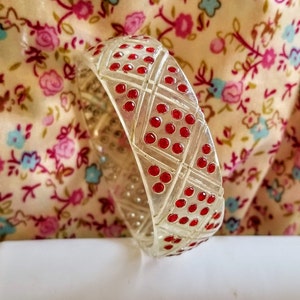 May include: A clear plastic bangle bracelet with a geometric design featuring red dots. The bracelet is a vintage style accessory.