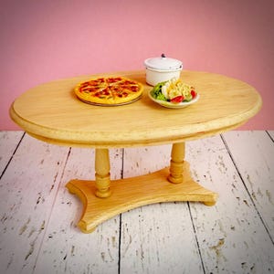 May include: A miniature wooden oval table with a light finish, holding a pizza, a salad, and a small white pot. The table is supported by four turned legs and a curved base. The background is a pink wall and a white-painted wooden floor.