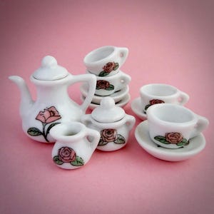 May include: A miniature white porcelain tea set with a teapot, creamer, sugar bowl, and several teacups and saucers. Each piece is decorated with a pink rose design. The set is arranged on a pink background.