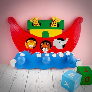 May include: A wooden Noah's Ark-themed wall hook, painted in bright colours. The red hull supports a yellow and green cabin, with a blue wave-shaped base. Features painted animal faces: lion, tiger, elephant, and giraffes. Includes three blue knobs.