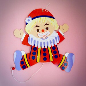 May include: A vintage clown cutout with a red hat, red and yellow outfit, and blue accents. The clown has a white ruffled collar, a painted smile, and a string for hanging. The background is a solid pink.