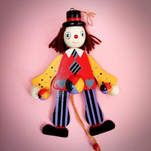May include: A wooden clown jumping jack toy featuring a black top hat, brown yarn hair, and a red nose. The clown wears a red vest with a diamond pattern, yellow sleeves with black dots, and purple striped pants. It holds small colorful balls.