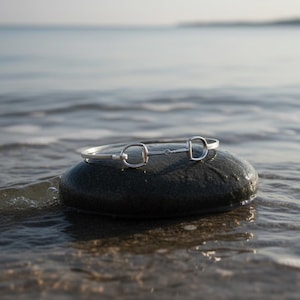 Könnte beinhalten: Ein silberfarbenes Armband im Equestrian-Stil mit einem Gebiss-Design liegt auf einem dunkelgrauen Stein. Das Armband wird am Wasserufer präsentiert, mit dem Meer im Hintergrund.