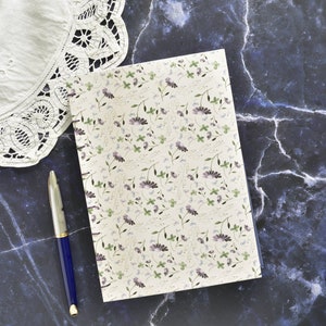 Op de afbeelding: Een spiraalgebonden notitieboek met een witte omslag en een bloemenpatroon in tinten van paars en groen. Een blauwe pen ligt op een donkerblauw marmeren oppervlak naast het notitieboek.