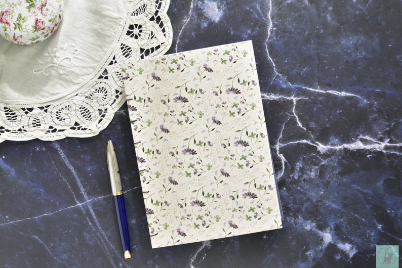 May include: A spiral bound notebook with a white cover and a floral pattern in shades of purple and green. A blue pen is lying on a dark blue marble surface next to the notebook.