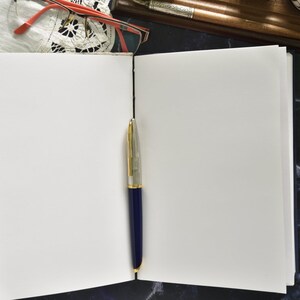 May include: An open blank notebook with a blue and gold pen lying on the open pages. The notebook is on a dark blue and white marble surface.