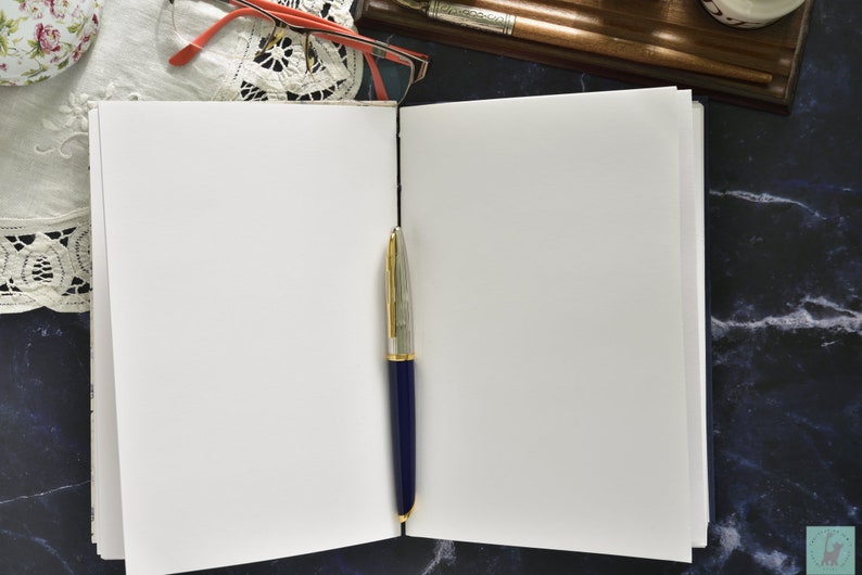 May include: An open blank notebook with a blue and gold pen lying on the open pages. The notebook is on a dark blue and white marble surface.