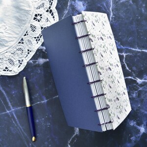May include: A handmade journal with a blue cover and white pages with a floral design. The journal is bound with purple thread and has a decorative spine.