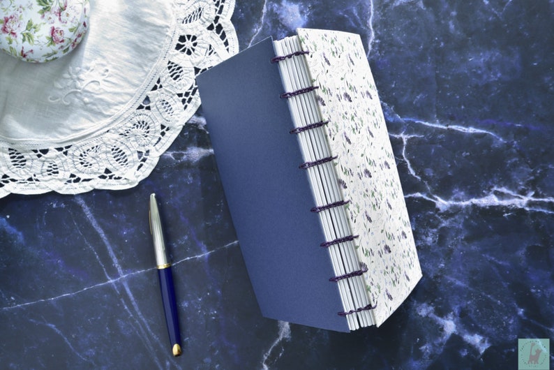 May include: A handmade journal with a blue cover and white pages with a floral design. The journal is bound with purple thread and has a decorative spine.