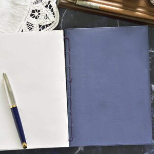 May include: A blank white notebook with a blue pen lying on top of it. The notebook is closed and has a blue cover. The notebook is on a black marble surface.