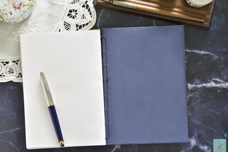 May include: A blank white notebook with a blue pen lying on top of it. The notebook is closed and has a blue cover. The notebook is on a black marble surface.