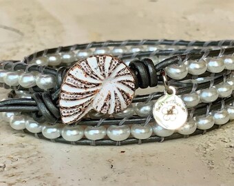 May include: Wrap bracelet featuring gray leather and white faux pearls. A white and brown seashell-shaped clasp and a small silver charm with a floral design add detail. The bracelet is designed to wrap around the wrist multiple times.