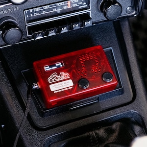 May include: A rectangular red translucent Melody Box electronic device featuring a white graphic of a motorcycle, two black circular buttons, and a small digital display screen. The device includes the text Melody Box and WWW.IMPORTBIBLE.COM.