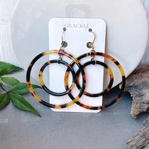 May include: A pair of large hoop earrings with a tortoiseshell pattern in shades of black, brown, and yellow. The earrings are double-hooped and hang from gold-colored hooks. The brand name "GRACKLE" is printed on the white product tag.