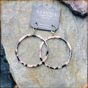 May include: Large circular hoop earrings featuring a mottled black, white, and translucent pattern. The earrings are attached to a dark gray display card with the text GRACKLE Handmade Jewelry Portland, OR and social media information.