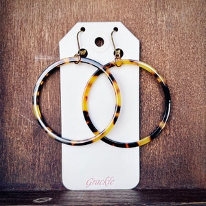May include: A pair of large, brown and yellow hoop earrings with gold-toned hardware. The earrings are displayed on a white card with the word "Grackle" printed on it.