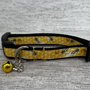 May include: A black cat collar with a yellow honeycomb pattern and bee accents. The collar has a silver metal ring with a gold bell and a bee charm attached. The collar is made of fabric and has a black plastic buckle.