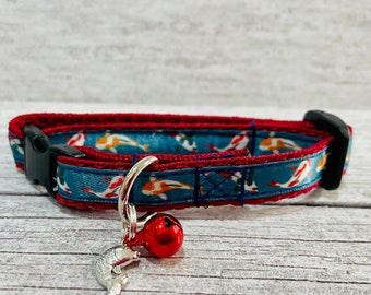 May include: A blue and red cat collar with a silver bell and a fish design. The collar has a buckle closure.