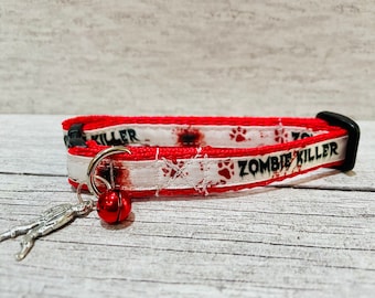 May include: Red and white cat collar with a bell and a zombie killer design. The collar has a black buckle and a silver ring.